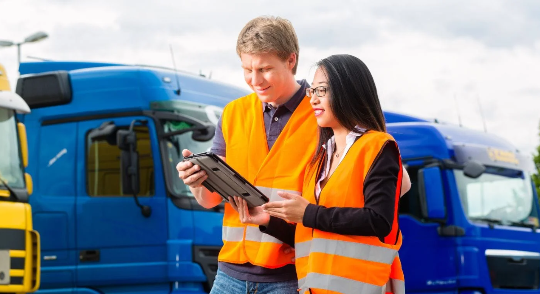 Dos trabajadores con chalecos reflectantes revisando datos en una tablet frente a camiones de transporte.