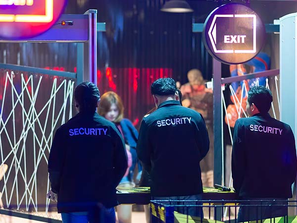 Tres agentes de seguridad controlando el acceso en la salida de un evento nocturno con detectores de metales y luces de neón.