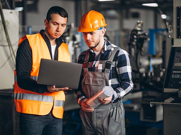 Dos trabajadores industriales revisando información en un portátil dentro de una fábrica; uno de ellos sostiene planos técnicos.